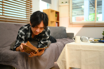 Happy young man lying on couch and reading book. Recreation and leisure activity concepts