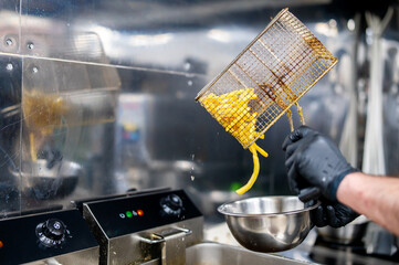 Hands in black gloves pouring fried potatoes from a metal fryer basket into a stainless steel bowl
