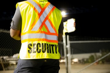 Back of a security guard patrolling a fenced commercial area at night.