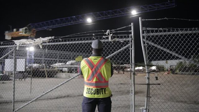 Back of a security guard patrolling a fenced commercial area at night.