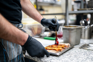 A person in a kitchen, wearing black gloves, applies sauce to a dish with a brush. The focus is on...