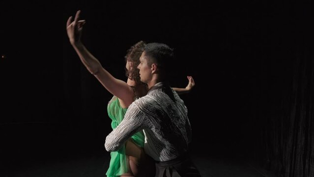 Ballroom couple dancing in darkness. Dancer in green chiton embraces her partner by neck standing with back to spectator. Man turns her by waist and pair performs support, girl on male shoulder sags