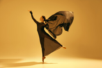 Beautiful young woman, ballet dancer in flowing black attire performing against golden color...