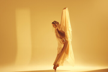 Dance of light. Artistic female ballet dancer in flowing golden dress making creative graceful performance against sand color background. Concept of art, classical dance, beauty, fashion, aesthetics