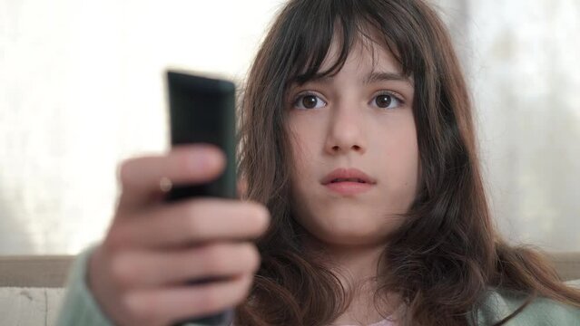 Child, teenage is sitting on the couch at home, using the remote control to switch channels on the TV.