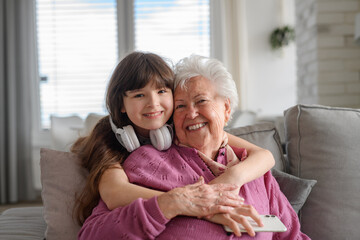 Cute girl hugging gradmother from behind, looking at camera, smiling. Portrait of an elderly woman...