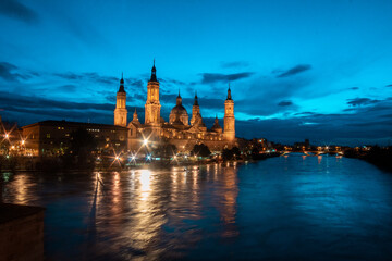 Fototapeta premium Twilight Majesty: Marveling at Zaragoza's Dusk, where the Iconic Basilica del Pilar, Stone Bridge, and Ebro River Unite in Spectacular Evening Splendor, Illuminated by the Fading Sunlight. 
