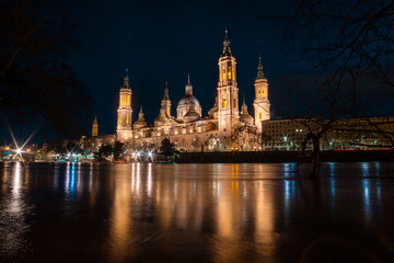 Naklejka premium Twilight Tranquility: Admiring the Serenity of Zaragoza's Evening Skyline, with the Iconic Basilica del Pilar and Stone Bridge Bathed in the Gentle Glow of Dusk, Overlooking the Meandering Ebro River.