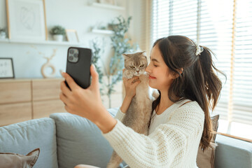 Smiling young asian woman taking selfie with Scottish fold cat on couch at home, Adorable domestic pet concept.