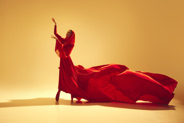 Passion in motion. Beautiful woman, dancer in flowing elegant red dress making powerful performance against sand color background. Concept of art, classical dance, beauty and fashion, aesthetics
