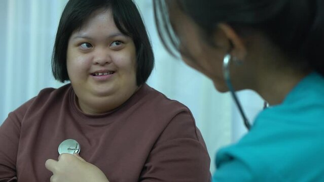 Asian female nurse uses stethoscope to perform physical examination of woman with Down syndrome at home.