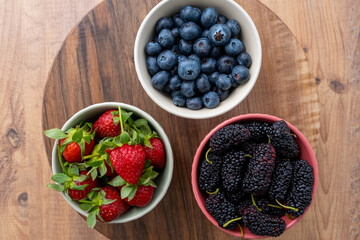 Top view of blueberries, black mulberries and strawberries in fruit cups on a wooden surface