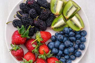 Close-up top view of blueberries, strawberries, blackberries and sliced kiwi fruit on a plate on a tablecloth