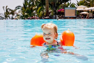 Little toddler girl with protective swimmies playing in outdoor swimming pool by sunset. Baby Child...