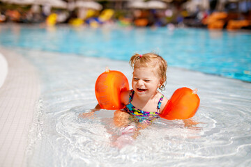 Little toddler girl with protective swimmies playing in outdoor swimming pool by sunset. Baby Child...