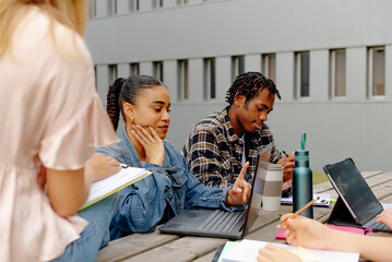 Fototapeta premium Multiracial group of young students studying or preparing a common class project, using laptops and sitting on a bench in the university campus gardens.