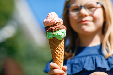 Happy preschool girl eating colorful ice cream in waffle cone on sunny summer day. Little toddler child eat icecream dessert. Sweet food on hot warm summertime days. Bright light, colorful ice-cream.