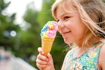 Happy preschool girl eating colorful ice cream in waffle cone on sunny summer day. Little toddler...