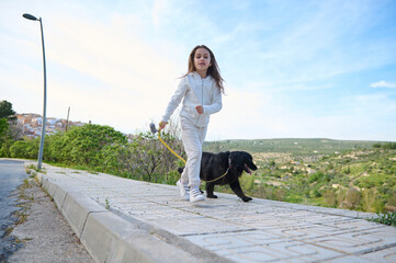 Happy child girl enjoying walking her dog outdoors in the nature. Pretty school age kid taking her...
