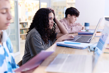 Side view portrait of teenage girl using laptop computer in library with multiethnic group of...