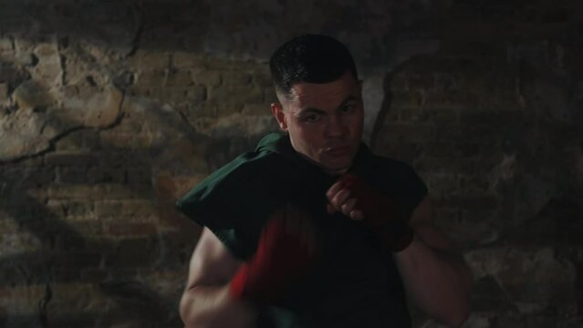 Focused young man in green T-shirt practicing his shadow boxing skills by throwing punch towards camera in dimly lit, rustic brick room, intense training and determination