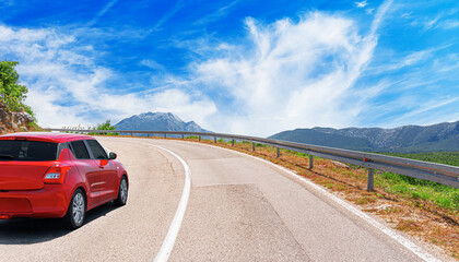 A red car drives along the highway against the backdrop of rocky mountains and tunnel on a sunny day.