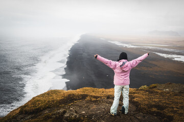 Young woman in pink jacket stand on top of mountain summit raises arms into air, happy and drunk on life, youth and happiness. Dyrholaey viewpoint tourist and wild atlantic ocean