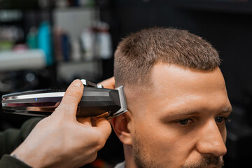 Skilled barber uses automatic trimmer to cut client hair on temple in barbershop closeup. Hairdresser does stylish hairdo by salon mirror