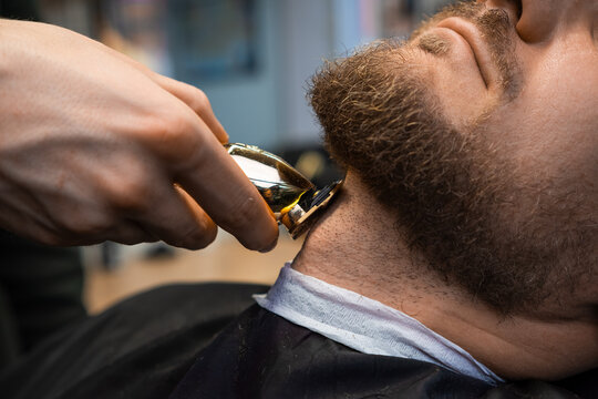 Stylist cuts hair on neck creating stylish beard shape for man with trimmer in barbershop closeup. Barber serves male client in beauty salon