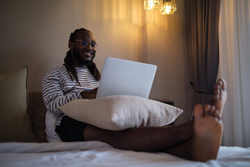 Smiling African American man using laptop on bed at home in the morning.