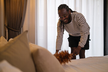 Young African American man doing house chores, cleaning up in bedroom. Household and cleaning...