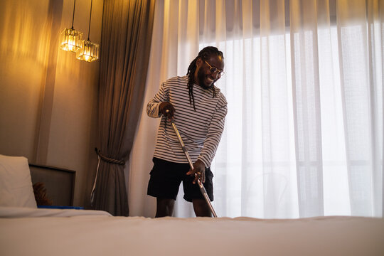 Young African American man doing house chores, cleaning up in bedroom. Household and cleaning concept.
