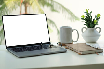 Laptop computer with blank screen, notepad and cup of coffee on white working desk near window.