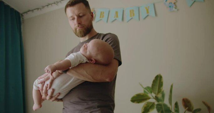 Young man dad holding infant baby on hands, rocking and putting child to sleep while standing at home in nursery room. Happy parenthood concept.