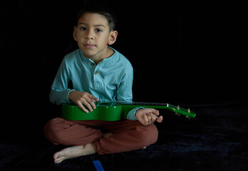 latin boy sitting cross-legged on the floor playing a green ukulele looking at the camera, black...