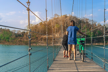 Back view of female tourist with son leads bicycle and enjoy nature across a high sling suspension...