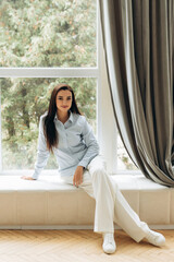 Stylish woman sitting on windowsill in front of big window. Portrait of businesswoman in formal style 