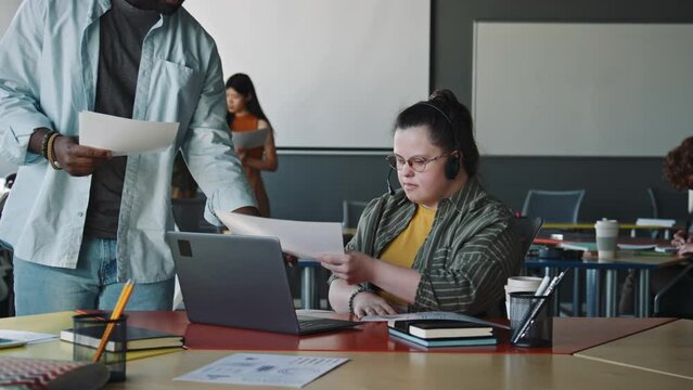 Young woman with Down syndrome in headset with mic speaking via web call on laptop and taking document from colleague while working as customer service manager in office