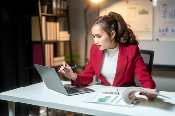 A woman in a red jacket is sitting at a desk with a laptop and a stack of papers. She is focused on her work and she is in a serious mood