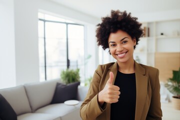 Portrait of a merry afro-american woman in her 30s showing a thumb up while standing against modern minimalist interior