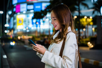 A portrait of a woman standing on the street at night, holding her smartphone and waiting for a ride.