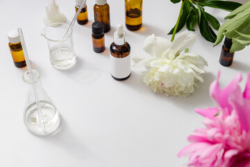 Lab glasswear, bottles of aroma oils, peony flowers on white desk. Empty space