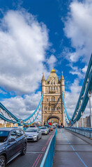 Obraz premium Car traffic and tourists walking along the street of Tower Bridge in London, with the view of the bridge's tensioning structures on a day with sunny blue sky and white clouds.