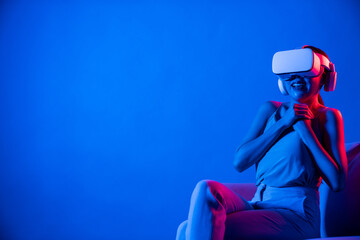 Smart female sitting on sofa surrounded by neon light wear VR headset connect metaverse, futuristic...