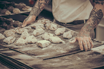 A baker with tattoos puts raw dough buns on the baking tray in the bakery