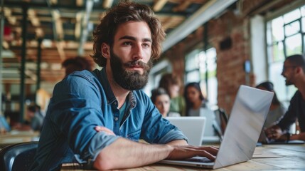 Motivated Caucasian young man with beard working on a laptop in an office. Male Director preparing marketing campaign. Diverse team working on computers.
