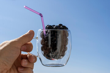 the hand holds a glass mug filled with blackcurrant berries with a cocktail tube