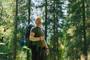 Thoughtful adult man with backpack trekking stick walking in green forest
