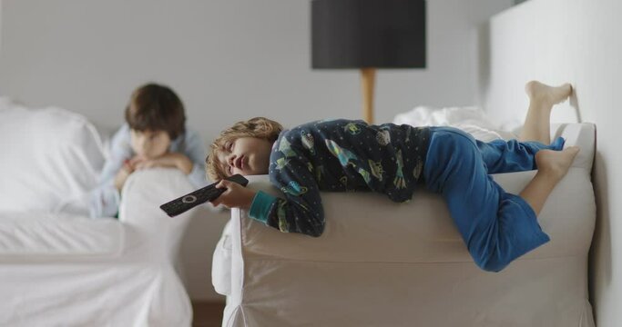 Two kids lounge idly in a living room, one lying on a couch with a remote in hand, eyes glued to the TV, the other sitting nearby, a picture of lethargy.