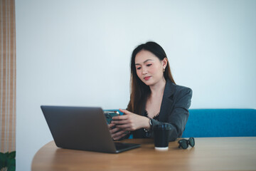 Asian young beautiful happy professional successful businesswoman designer sitting smiling at workstation desk using laptop notebook computer and smartphone working remotely online at home office